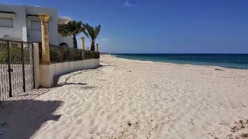 A sandy beach with a white building and palm trees in the background.