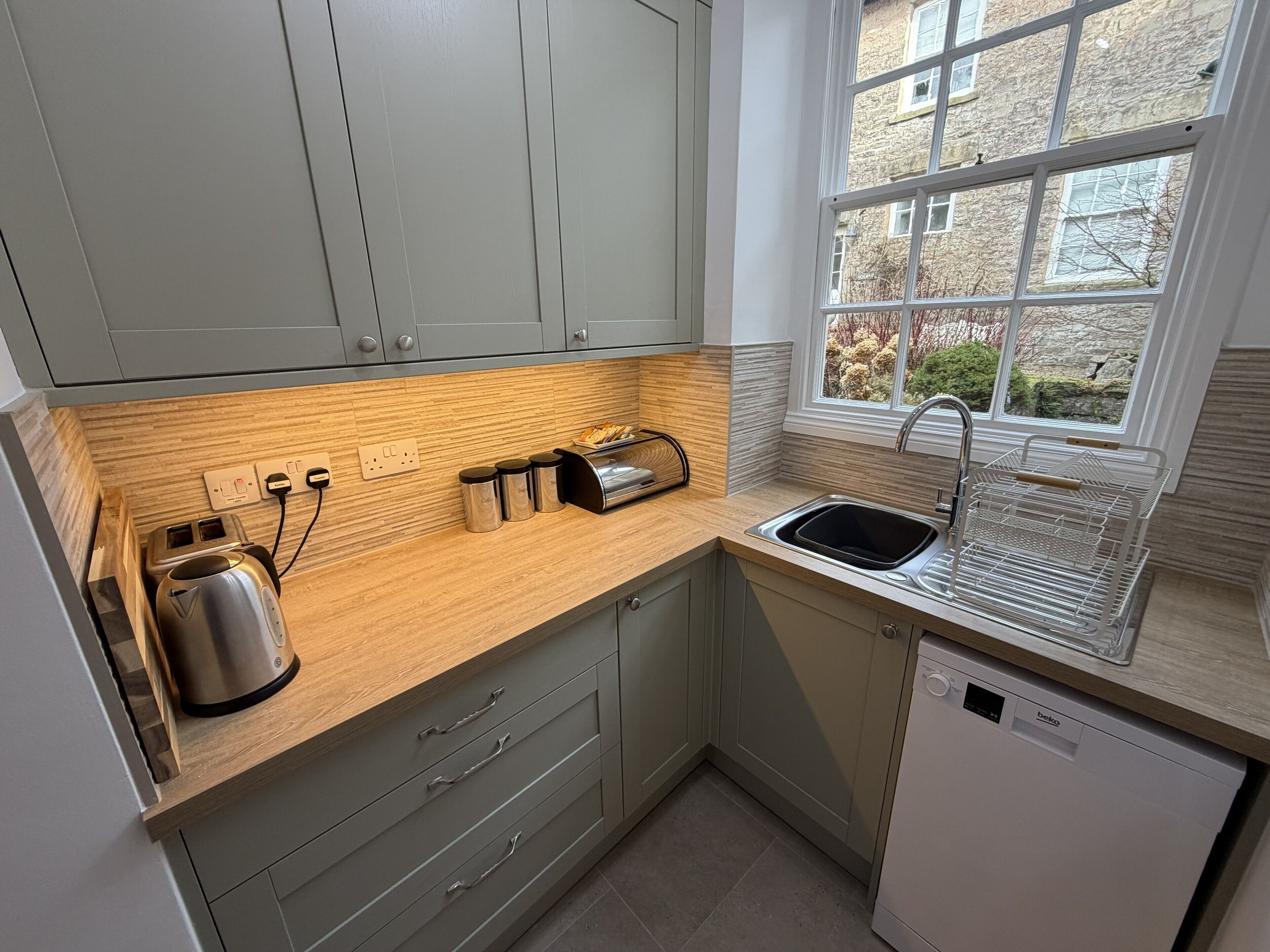 Een moderne keuken met grijze kastjes, een houten aanrechtblad, een inbouw vaatwasser en een raam met uitzicht op gebouwen.