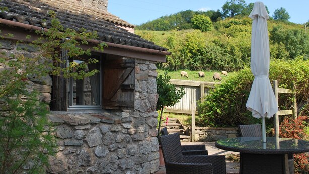 A stone building with a tiled roof, a wooden door, and a small outdoor seating area with wicker chairs and a table.