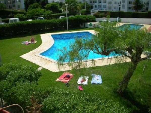 A swimming pool surrounded by a grassy area with a tree and a building in the background. - Apartment (Albufeira)