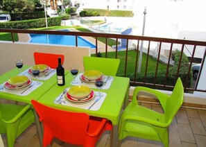 A balcony with a pool view, green and red chairs, and a table set for two with plates, glasses, and a bottle.