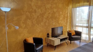 Ett modernt vardagsrum med en platt-TV på ett träställ, två svarta läderfåtöljer med orange kuddar, en golvlampa och ett stort fönster med tunna, genomskinliga gardiner.
