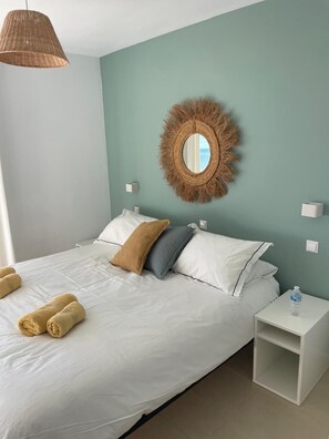 A neatly made bed with white linens and rolled towels, a round mirror with a fringe border, a white nightstand with a water bottle, and a hanging light fixture.