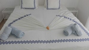 A neatly made bed with a white bedspread and blue zigzag trim, two pillows with similar trim, and two rolled towels placed at the foot of the bed.