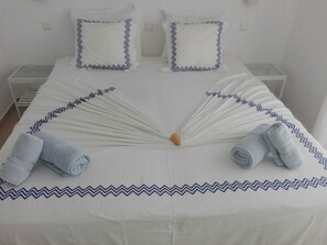 Un lit bien fait, recouvert d’une courtepointe blanche ornée d’un motif en zigzag bleu, avec deux oreillers assortis et deux serviettes enroulées, posées au pied du lit.