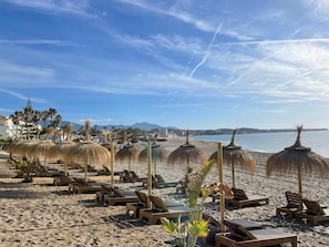 Une plage bordée de rangées de chaises longues et d’ombrelles en chaume, un ciel dégagé et une mer calme.