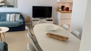 A modern living space with a sofa, a flat-screen TV, a dining table with chairs, and a kitchen area with a coffee maker.