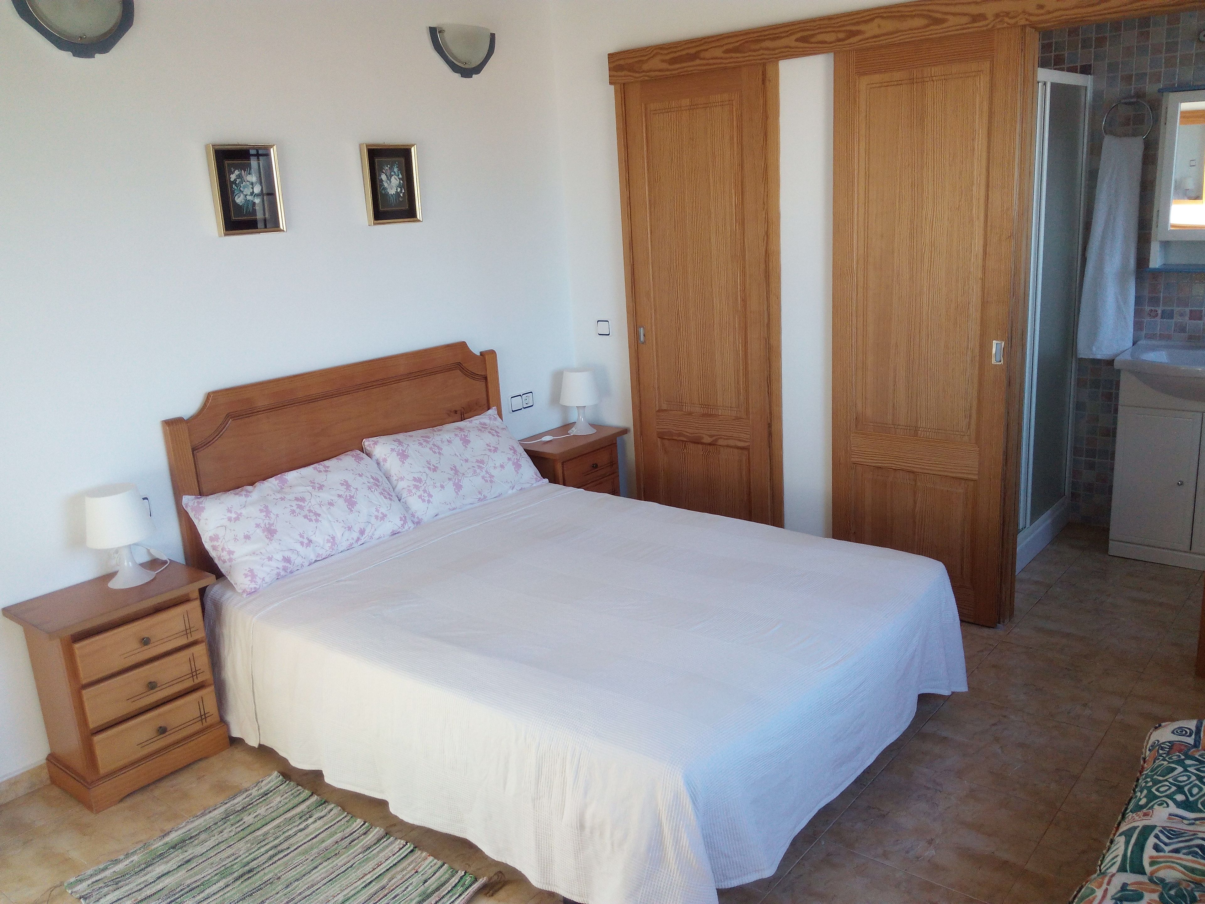 Una cama bien hecha con cabecera de madera, una mesita de noche con lámpara y un cajón pequeño. Dos cuadros enmarcados están colgados en la pared sobre la cama.