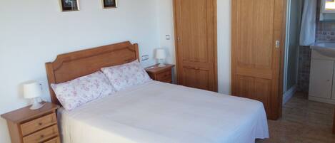 Una cama bien hecha con cabecera de madera, una mesita de noche con lámpara y un cajón pequeño. Dos cuadros enmarcados están colgados en la pared sobre la cama.