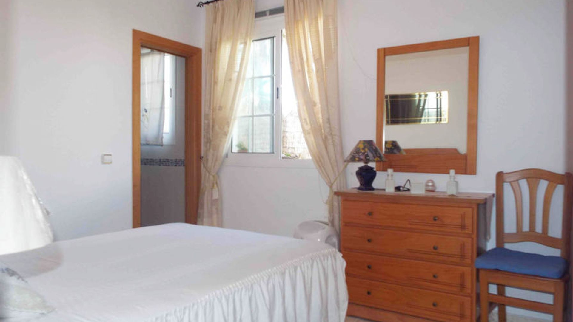 A bedroom with a bed, a wooden dresser, a chair, a mirror, and a window with curtains.