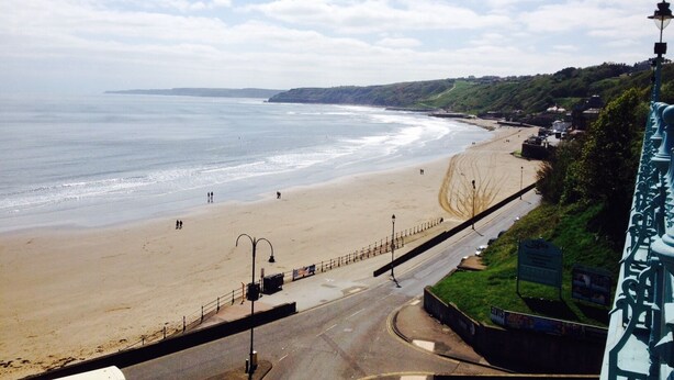 A coastal road with a beach on one side and a building with a balcony on the other.
