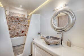 A bathroom with a stone wall, a bathtub, a round mirror, and a stone sink.