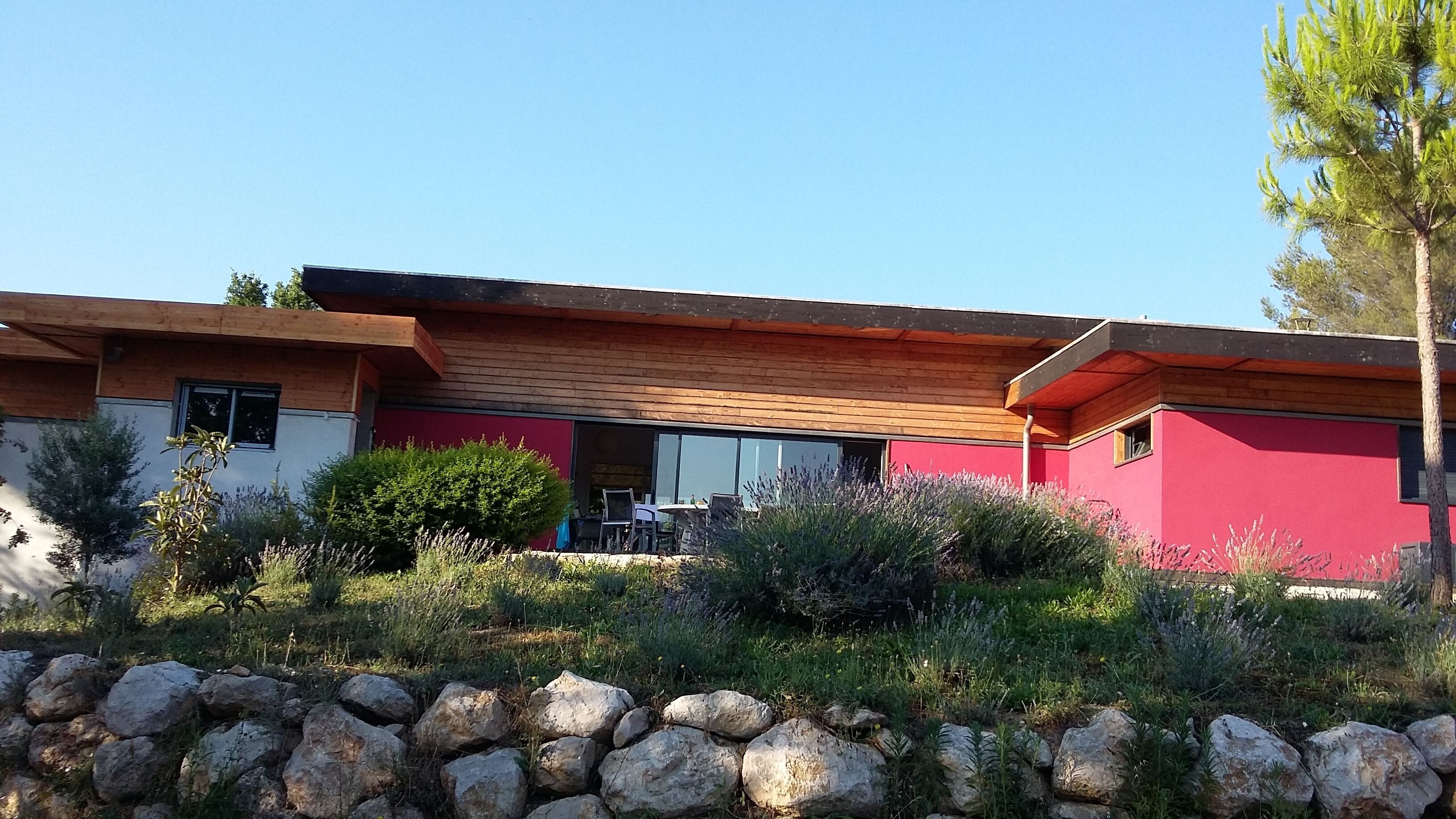 Une maison moderne avec une façade rouge, un toit en bois et un mur de pierre au premier plan.