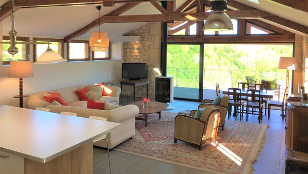 A spacious living area with a high ceiling, wooden beams, a fireplace, a television, and a dining area with outdoor views. - Luxury renovation of an ancient stone barn in a medieval bastide village (Montcabrier)