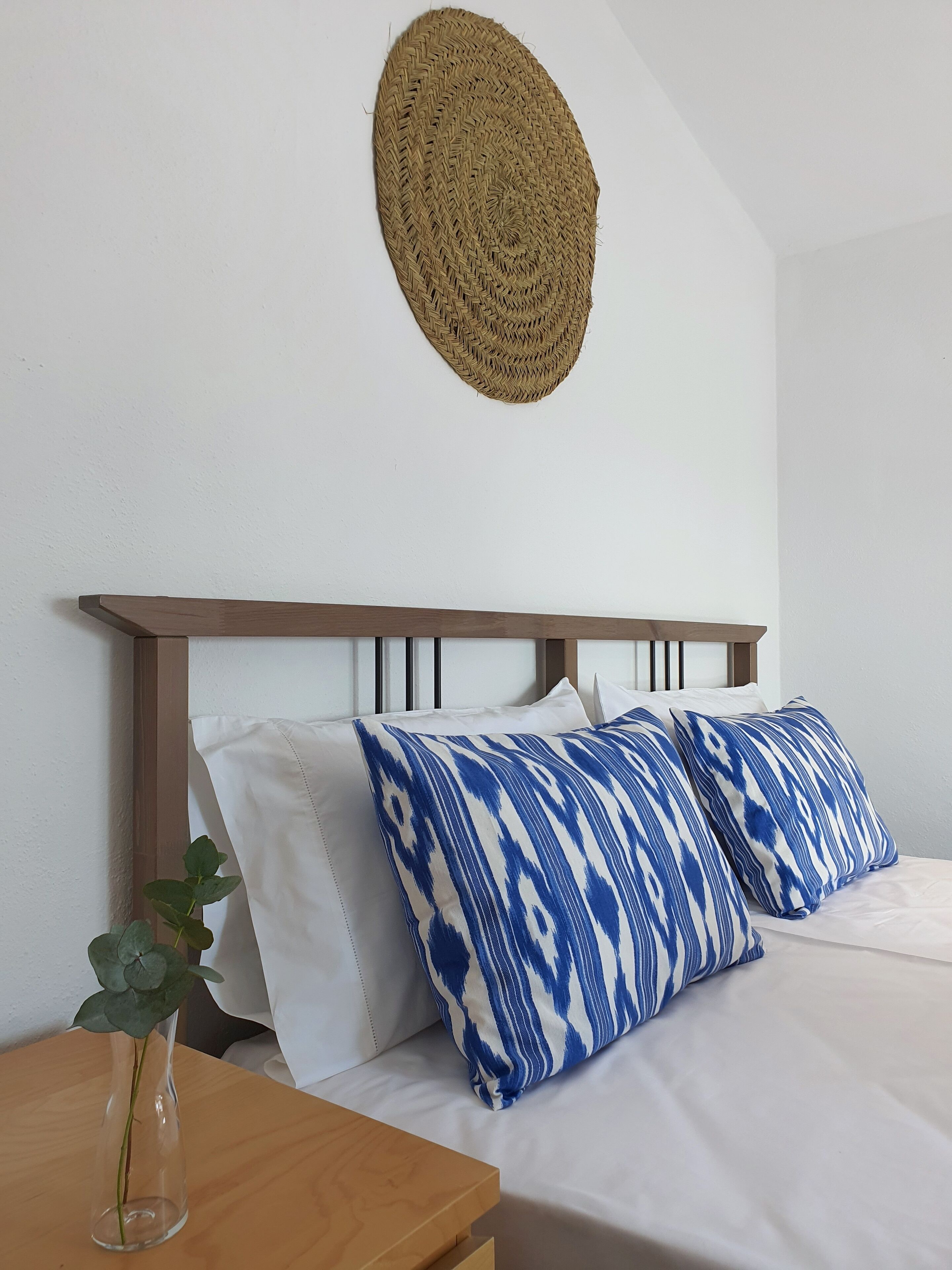Un lit bien fait, agrémenté d’oreillers blancs et bleus à motifs, un vase transparent contenant une seule plante verte, et une décoration murale tissée au-dessus du lit.