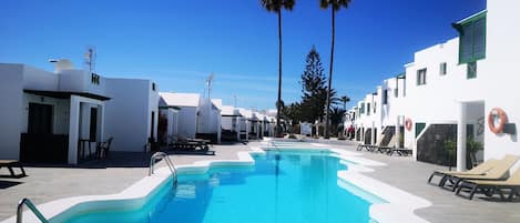 A swimming pool with clear blue water, surrounded by white buildings with green shutters, and palm trees in the background.