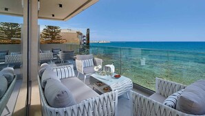 A balcony with white wicker furniture, a table with a vase and a bowl of fruit, and a clear view of the sea and distant buildings. - Seafront Apt, beachfront, quiet & central with infinity pool (Limenas Chersonisou)