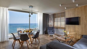 A modern living room with a large window overlooking the sea, a dining area with a table set for two, and a comfortable seating area with a sofa and armchairs. - Seafront Apt, beachfront, quiet & central with infinity pool (Limenas Chersonisou)