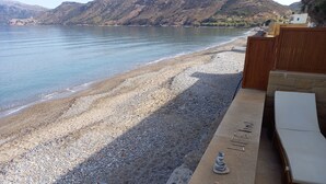 A beach with a pebble shore, a wooden fence, and a lounge area with a stone wall and a small statue.