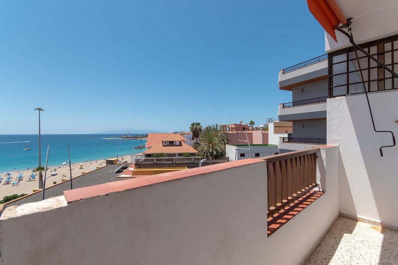 A coastal view with a beach, a building with a red-tiled roof, and a balcony with a railing.