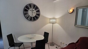 Ein Zimmer mit einem runden Tisch, vier Stühlen, einer großen Wanduhr, einer Stehlampe, einem roten Sofa und einem gerahmten Spiegel.