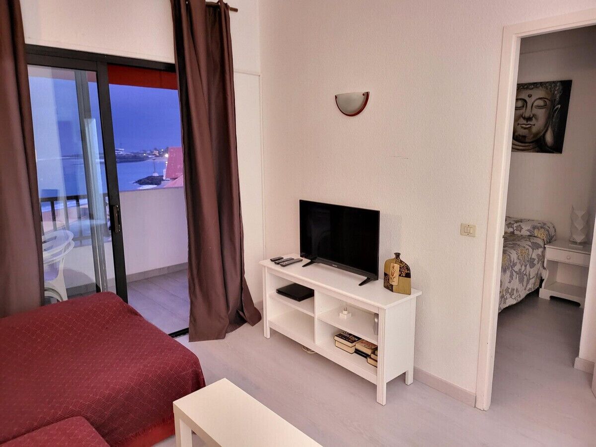 Ein Hotelzimmer mit Bett, Fernseher auf einem weißen Schrank, einem kleinen Tisch und Meerblick durch eine Glastür.