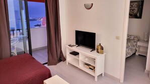 Ein Hotelzimmer mit Bett, Fernseher auf einem weißen Schrank, einem kleinen Tisch und Meerblick durch eine Glastür.