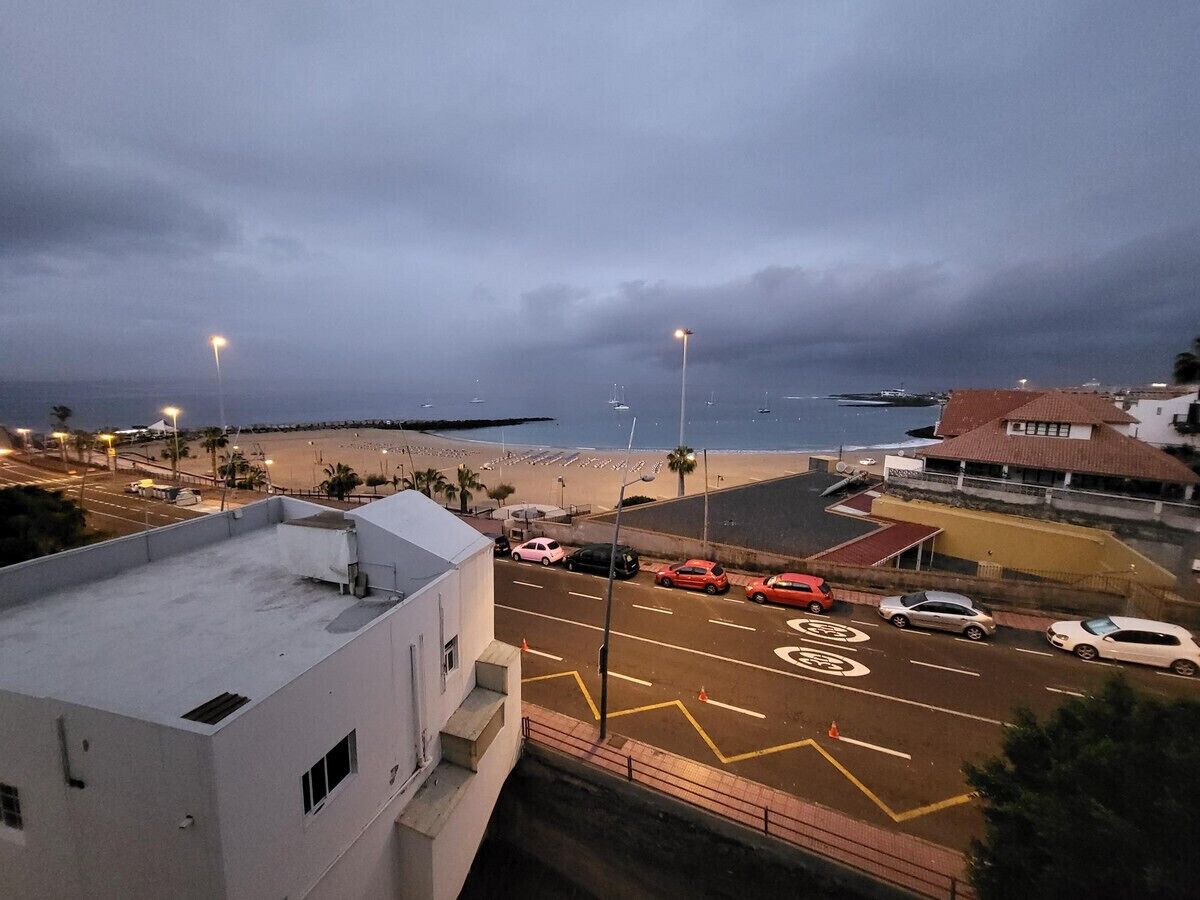 A coastal road with cars parked on the side, a beach in the background, and a building with a flat roof in the foreground.