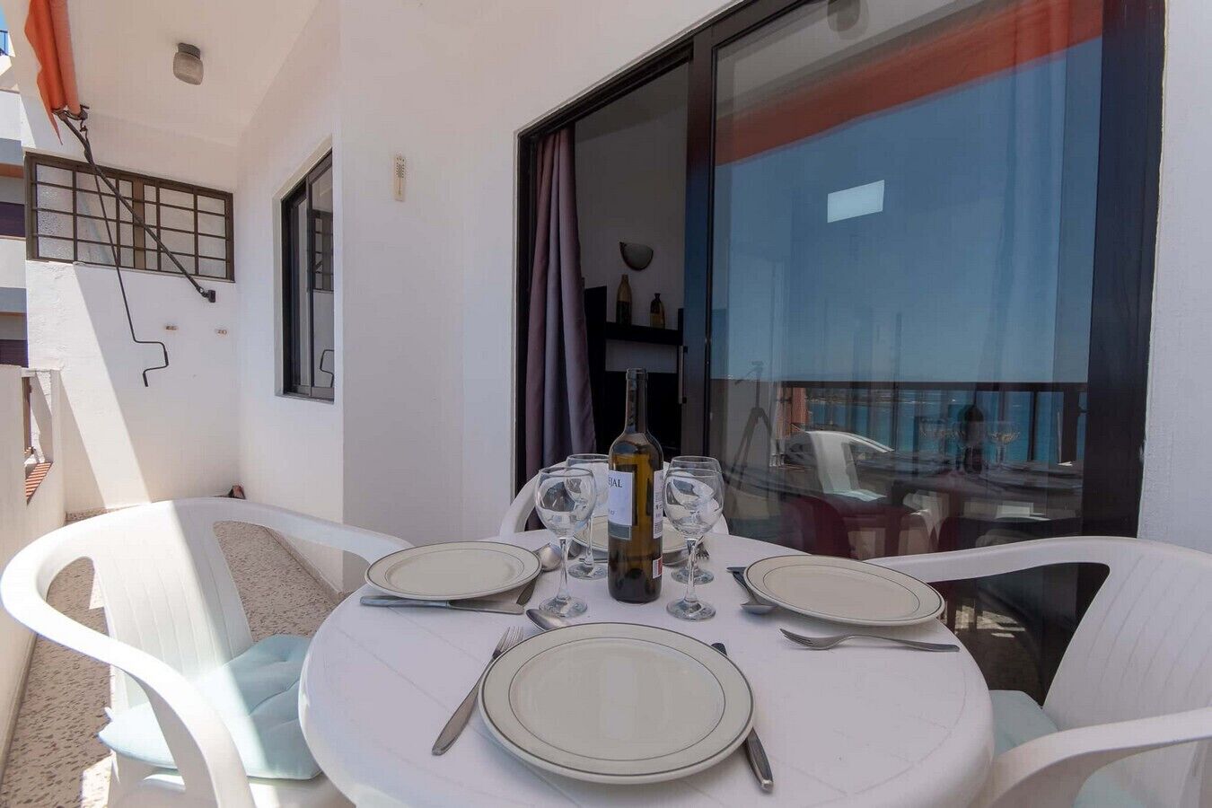Uma mesa de jantar redonda posta com pratos, talheres e uma garrafa de vinho, rodeada por cadeiras brancas numa varanda com vista para o mar.