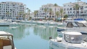 Un puerto deportivo con varias embarcaciones amarradas, rodeado de edificios de varios pisos con balcones y palmeras.