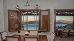 A dining area with a wooden table and chairs, a vase of flowers, and a view of the sea through open windows. - Villa Imperia 3 bedroom luxury villa - 200m from the beach (Elounda)