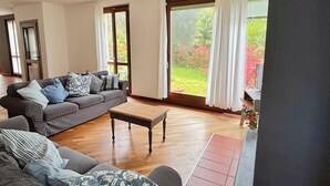 Un salon spacieux avec un canapé gris, une table basse en bois et de grandes fenêtres donnant sur un jardin.