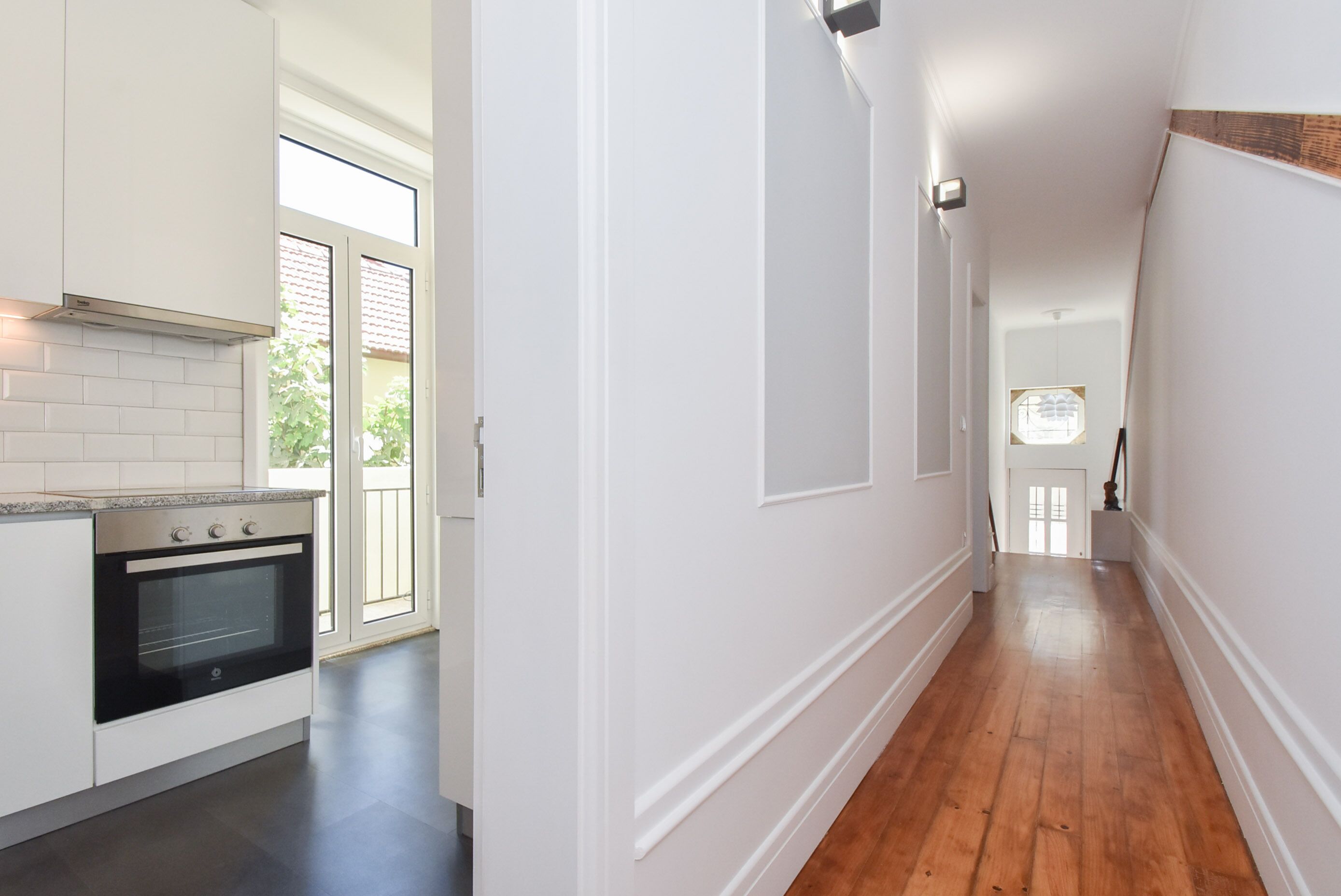 A narrow hallway with wooden flooring and white walls, leading to a kitchen area with a modern stove and white cabinets.