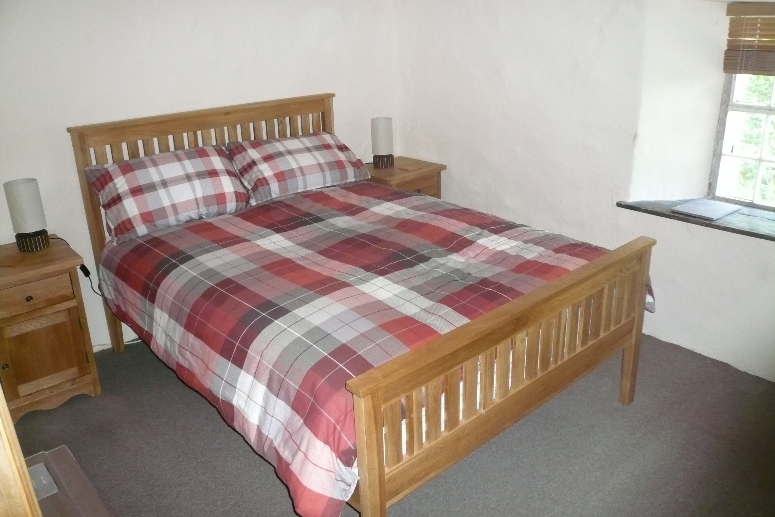 A single-bed room with a wooden headboard, a plaid bedspread, two bedside tables with lamps, and a window with blinds.