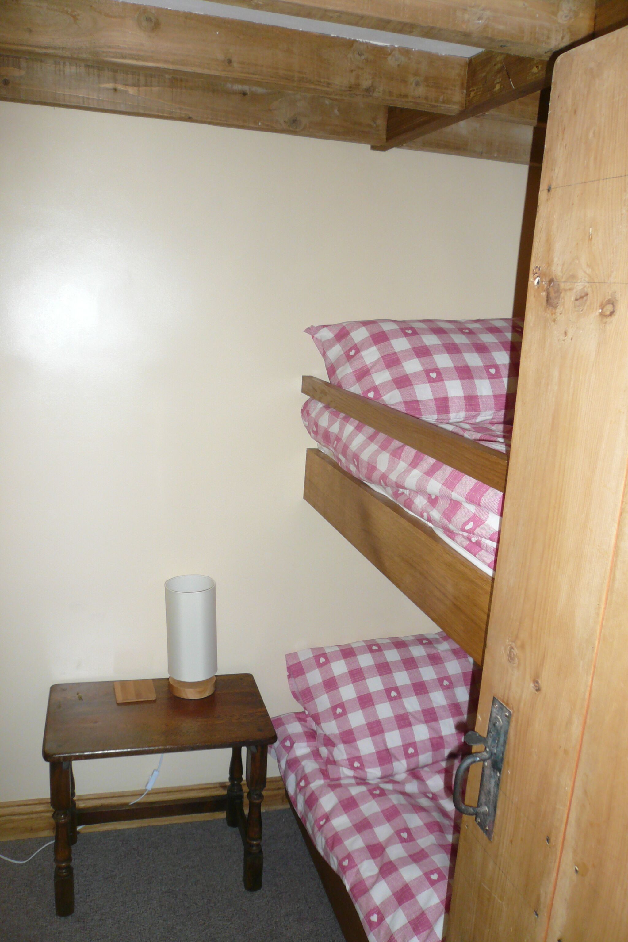 Un lit superposé avec une literie à carreaux, une petite table en bois sur laquelle se trouve un objet cylindrique, et une porte en bois.