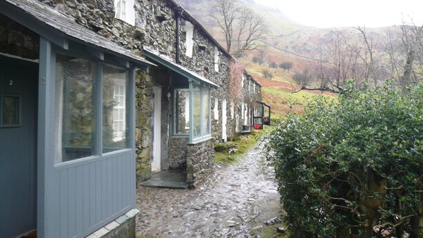 A stone building with a porch and large windows, set in a rural landscape with a stone pathway and greenery. - Cosy, romantic fellside cottage hidden in the Glencoyne valley (Glenridding)