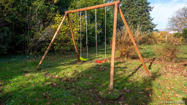 Ein hölzernes Schaukelgerüst in einer grünen Wiese mit Bäumen im Hintergrund.