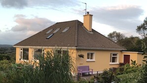 Een geel huis met een schuin dak, schoorsteen en een tuin met een bank.