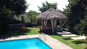 A swimming pool with a thatched gazebo, lounge chairs, and a stone wall.