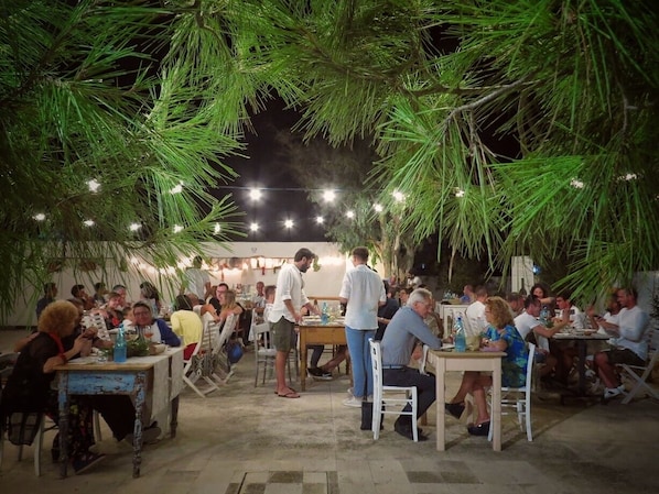 An outdoor dining area with tables and chairs, surrounded by greenery and string lights.