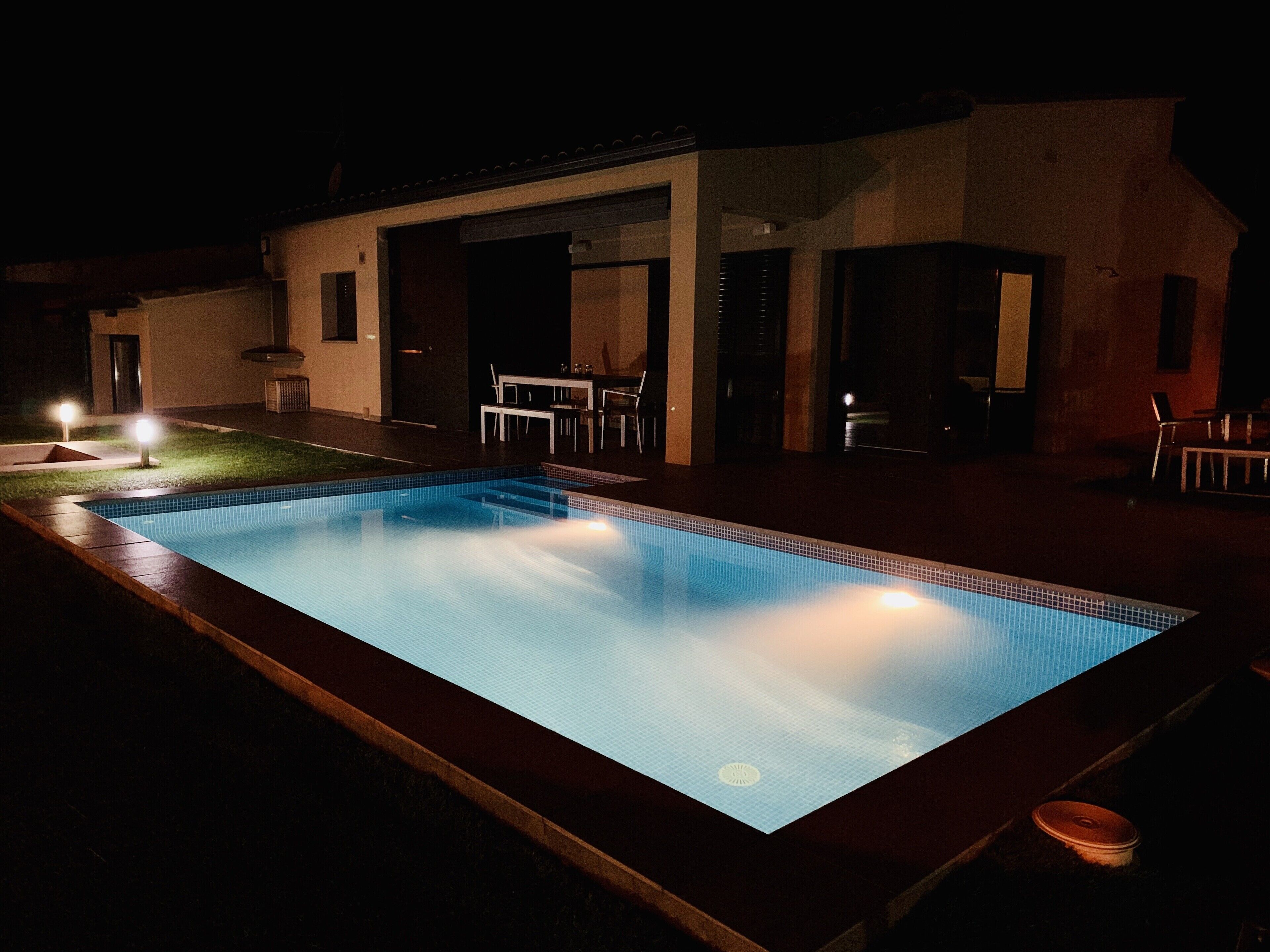 A rectangular swimming pool with a tiled edge, surrounded by a deck. There is a covered outdoor seating area with tables and chairs.