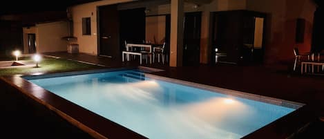 En rektangulär swimmingpool med en kant som är klädd med kakel, omgiven av en terrass. Det finns ett takförsett utomhusområde med bord och stolar där man kan sitta.
