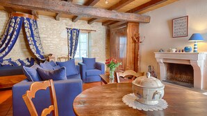 Una sala de estar acogedora con pared de piedra, vigas de madera, mobiliario azul, chimenea y un jarrón con flores.