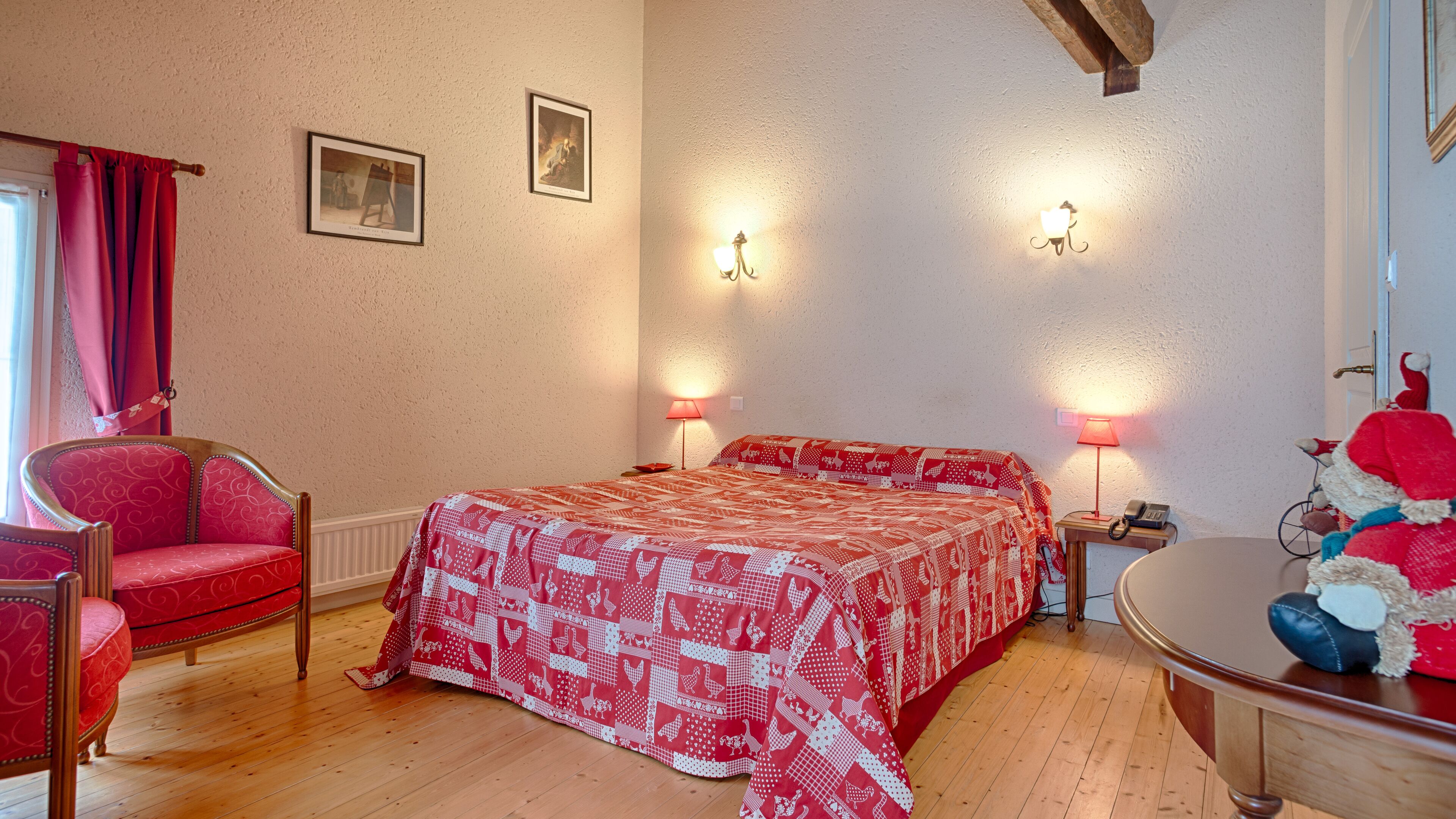 A bedroom with a wooden floor, a red patterned bedspread, two wall-mounted lamps, a chair, and a small table with a lamp.