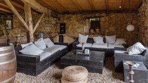 A rustic stone-walled room with a wooden ceiling, furnished with black sofas and a coffee table, and a woven basket on the floor. - CHARMING VILLAGE HOUSE! near Bergerac StEmillion Bordeaux, heated pool jacuzzi (Saint-Martin-De-Gurson)