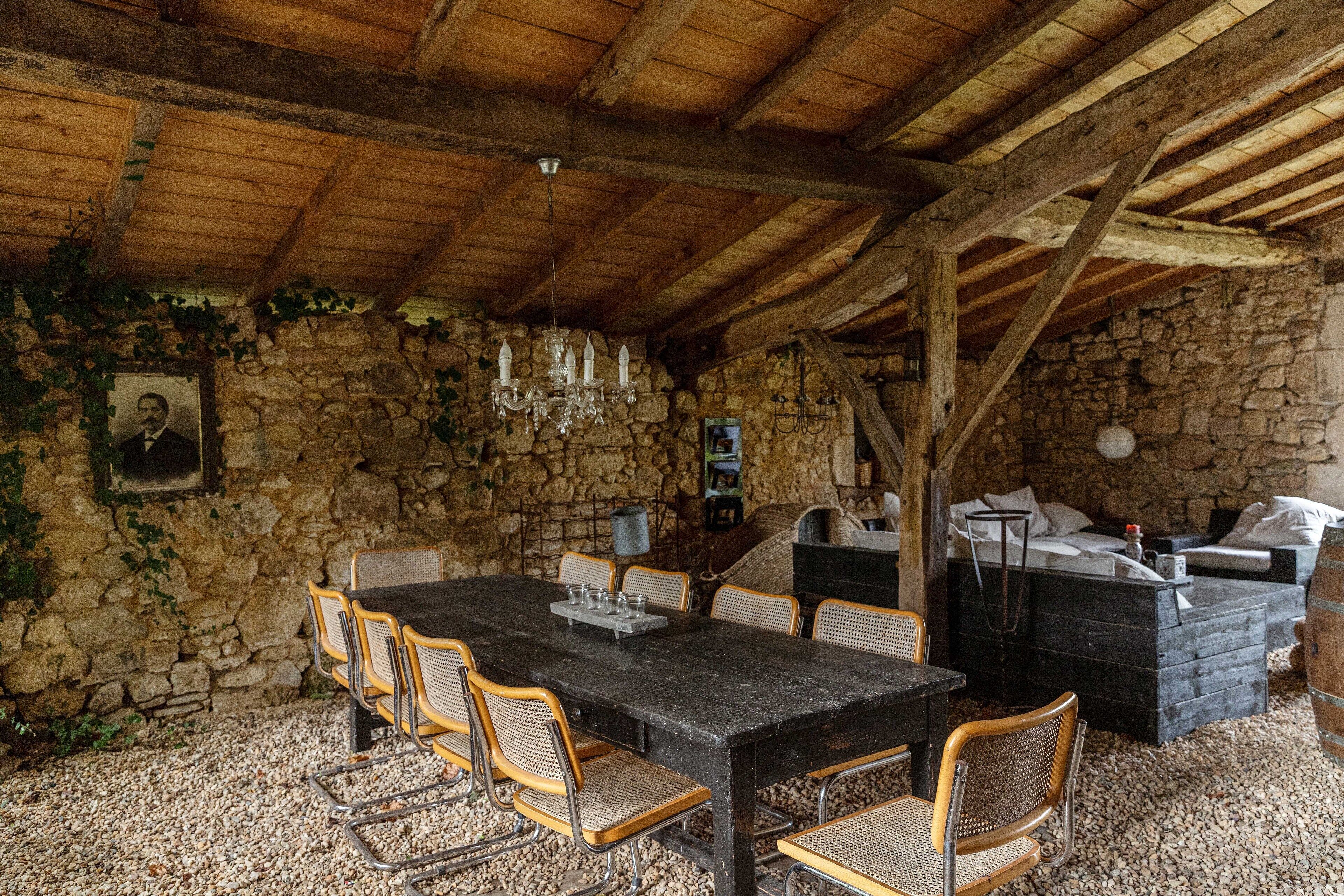 A rustic stone-walled room with a wooden ceiling, a dining table with chairs, a sofa, and a small bed.