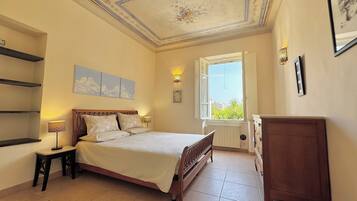 Une chambre à coucher comprenant un lit, une table de chevet, une commode, une fenêtre avec un volet ouvert et un plafond orné de motifs floraux.