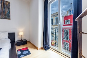 A bedroom with a bed, bedside table, lamp, and a view of a red building through a window.