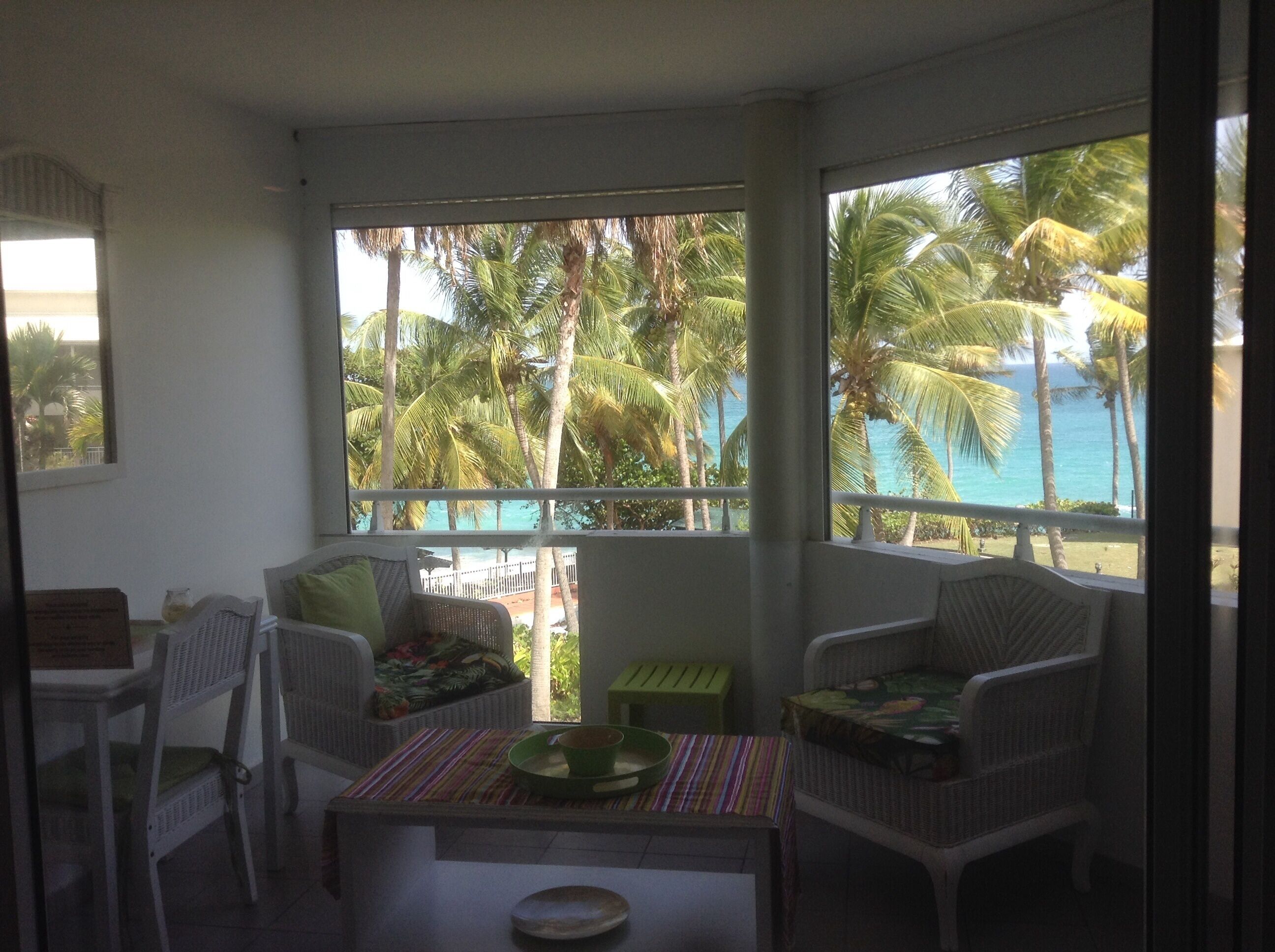 A balcony with wicker furniture, a small table, and a view of palm trees and the ocean.