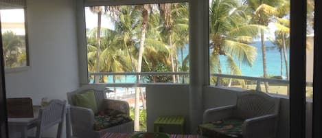 A balcony with wicker furniture, a small table, and a view of palm trees and the ocean.